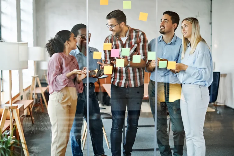 Cinq jeunes collègues discutent devant une paroi vitrée couverte de post-it colorés